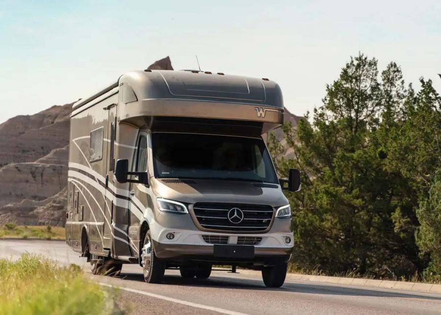 2023 Winnebago Navion driving on a roadway with rocky hill behind it