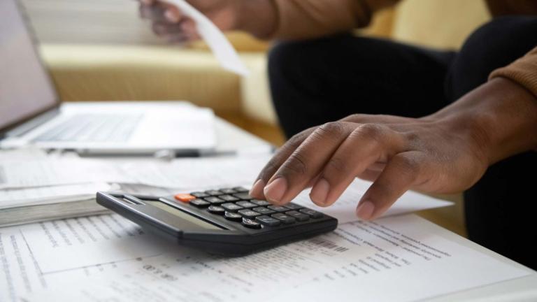 Man's hand above a calculator with paperwork and a computer in the background