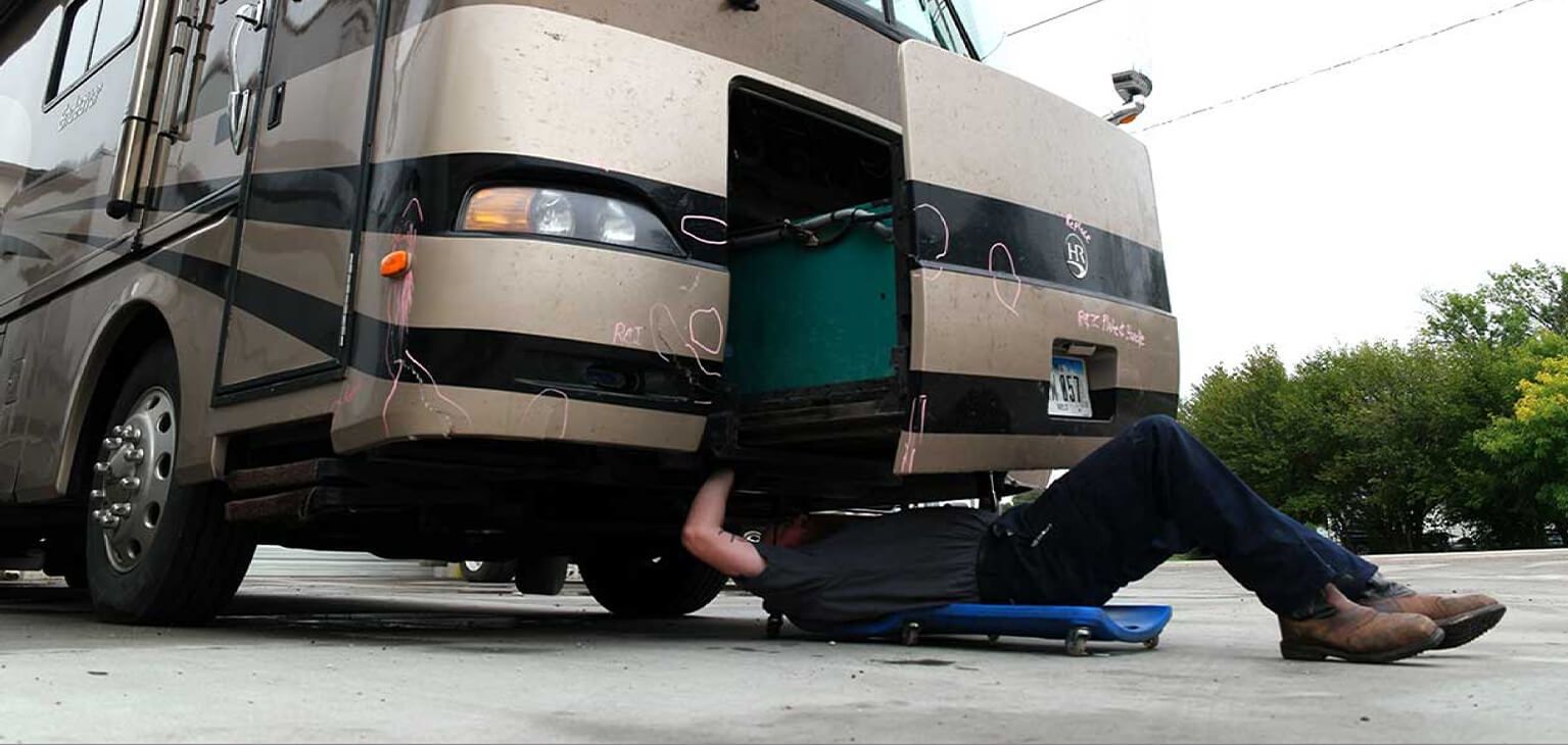 A mechanic in dark clothing and boots works on the underside of a gold RV with black stripes.